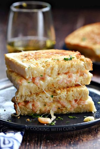 A grilled cheese sandwich with melted cheese and shrimp filling, garnished with parsley on a dark plate, accompanied by grilled cheese sandwiches, with a glass of wine in the background.