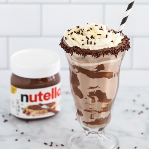 An easy Nutella milkshake served in a glass with whipped cream, chocolate sprinkles, and a straw, sits next to a jar of Nutella on a marble surface.