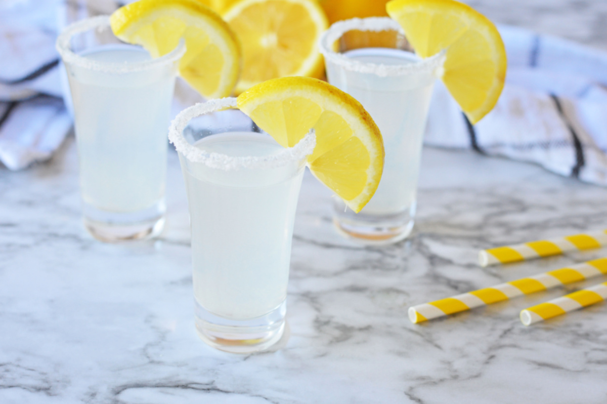 Three glasses with lemon slices and straws on a marble table, showcasing a refreshing lemon drop shot recipe.