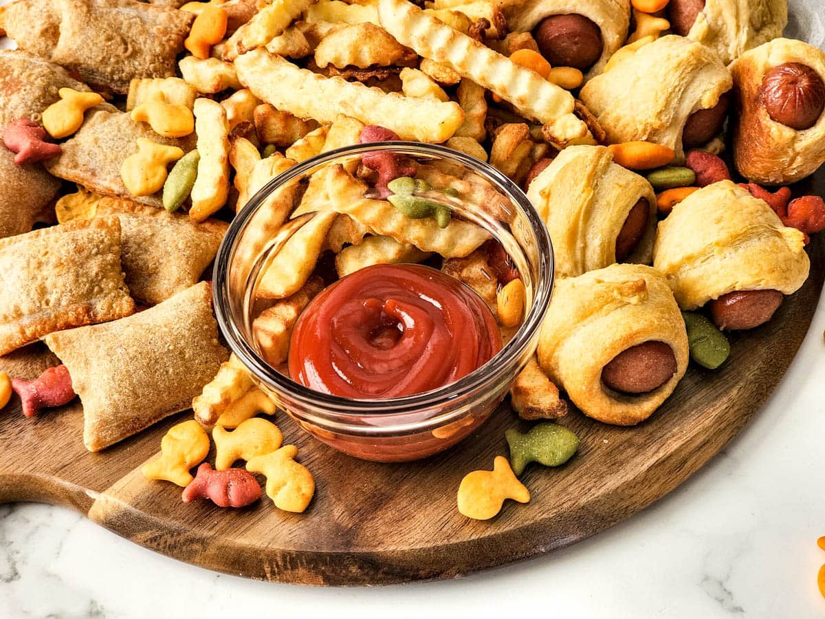 Bowl of ketchup surrounded by pizza rolls, fries and pigs in a blanket