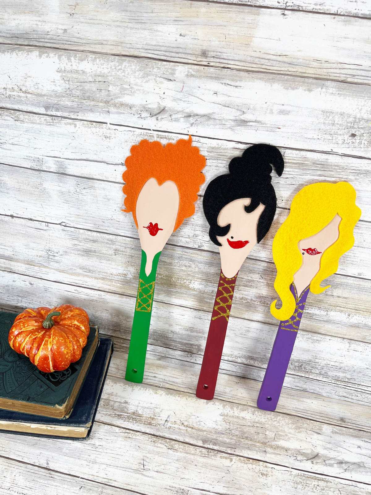 Three wooden spoon witches on a table next to pumpkins.
