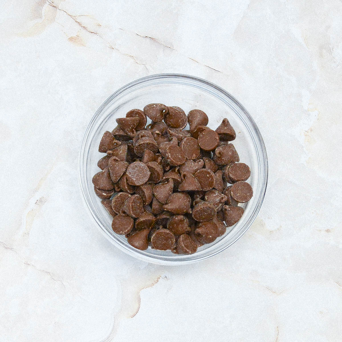 Chocolate chips in a glass bowl on a marble surface.