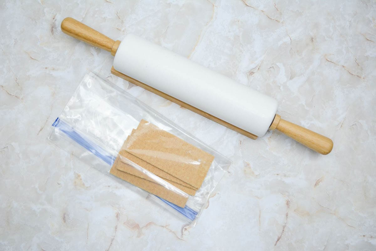 A rolling pin and a plastic bag with graham crackers on a marble table.