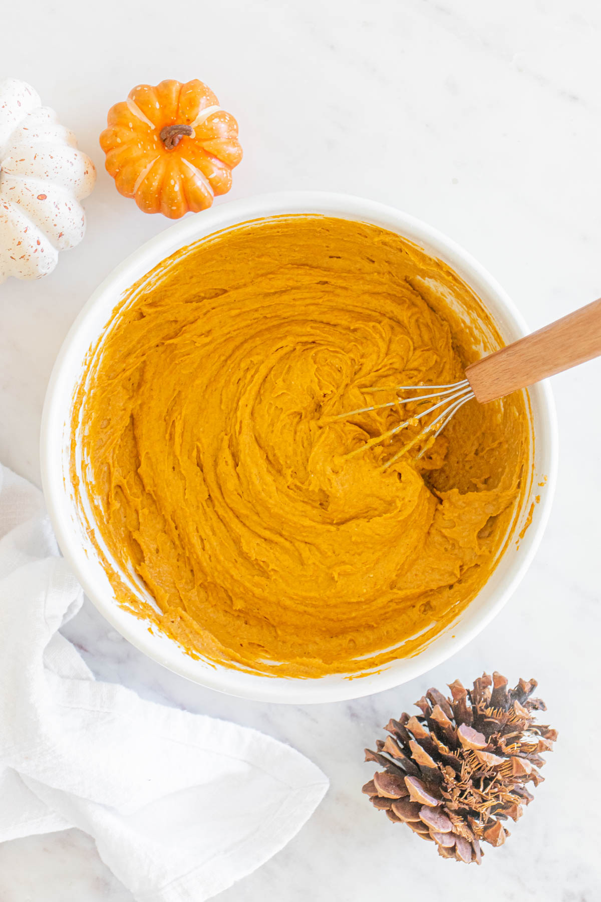 A bowl of pumpkin cookie dough with a whisk and pine cones next to it.