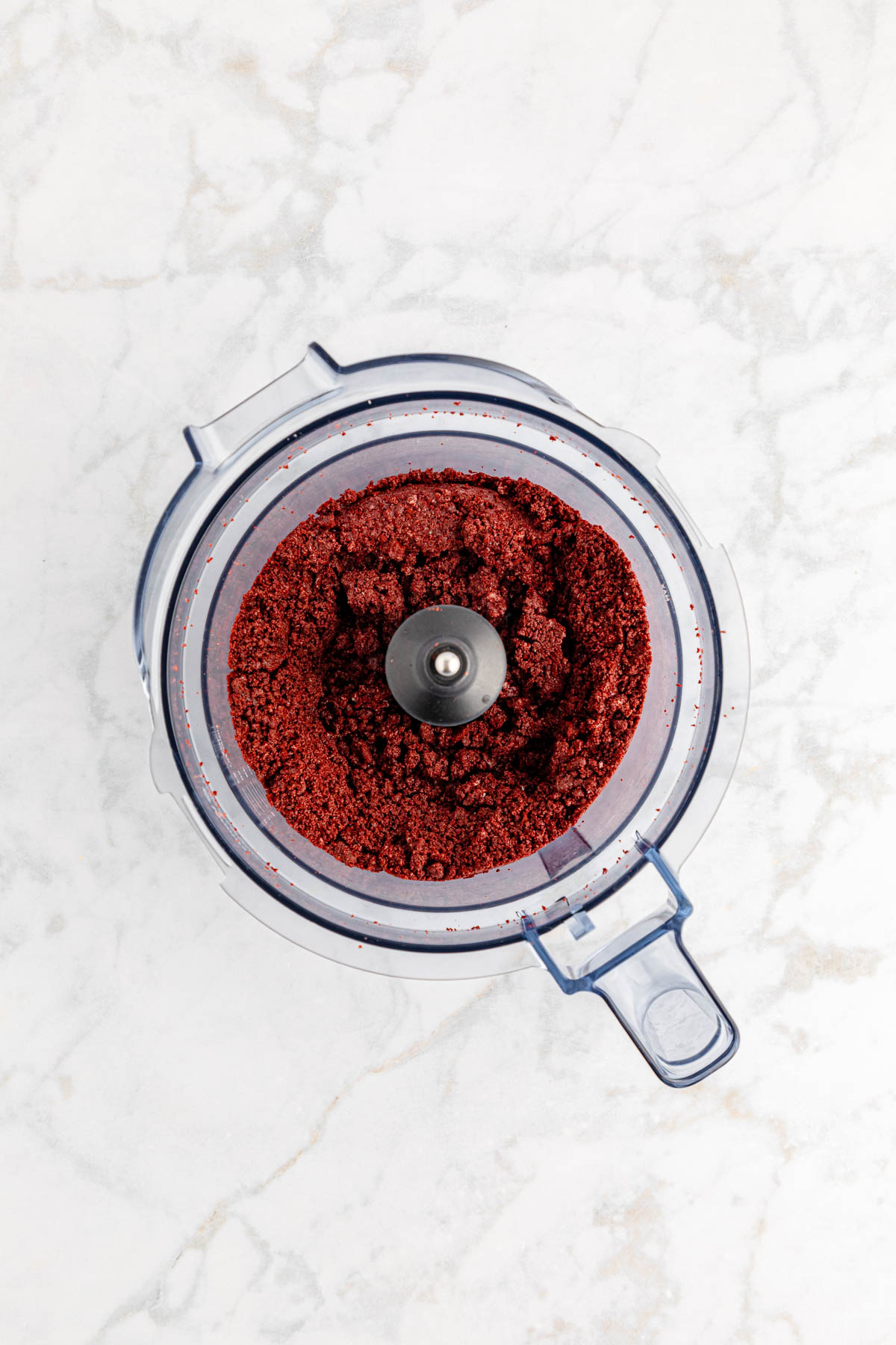 Red velvet oreo cookie crumbs in food processor on a marble surface.