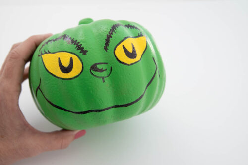 A person is holding up a green pumpkin painted with the grinch's face.