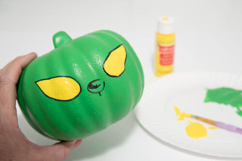 A person painting a green pumpkin with yellow eyes.