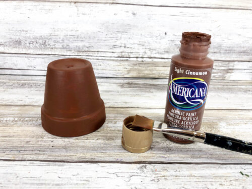 A bottle of brown paint next to a paint brush and clay pot painted brown