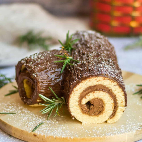A chocolate roll with rosemary sprigs on a wooden cutting board.