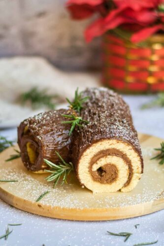 A chocolate Christmas roll displayed on a wooden cutting board amongst other festive cakes.