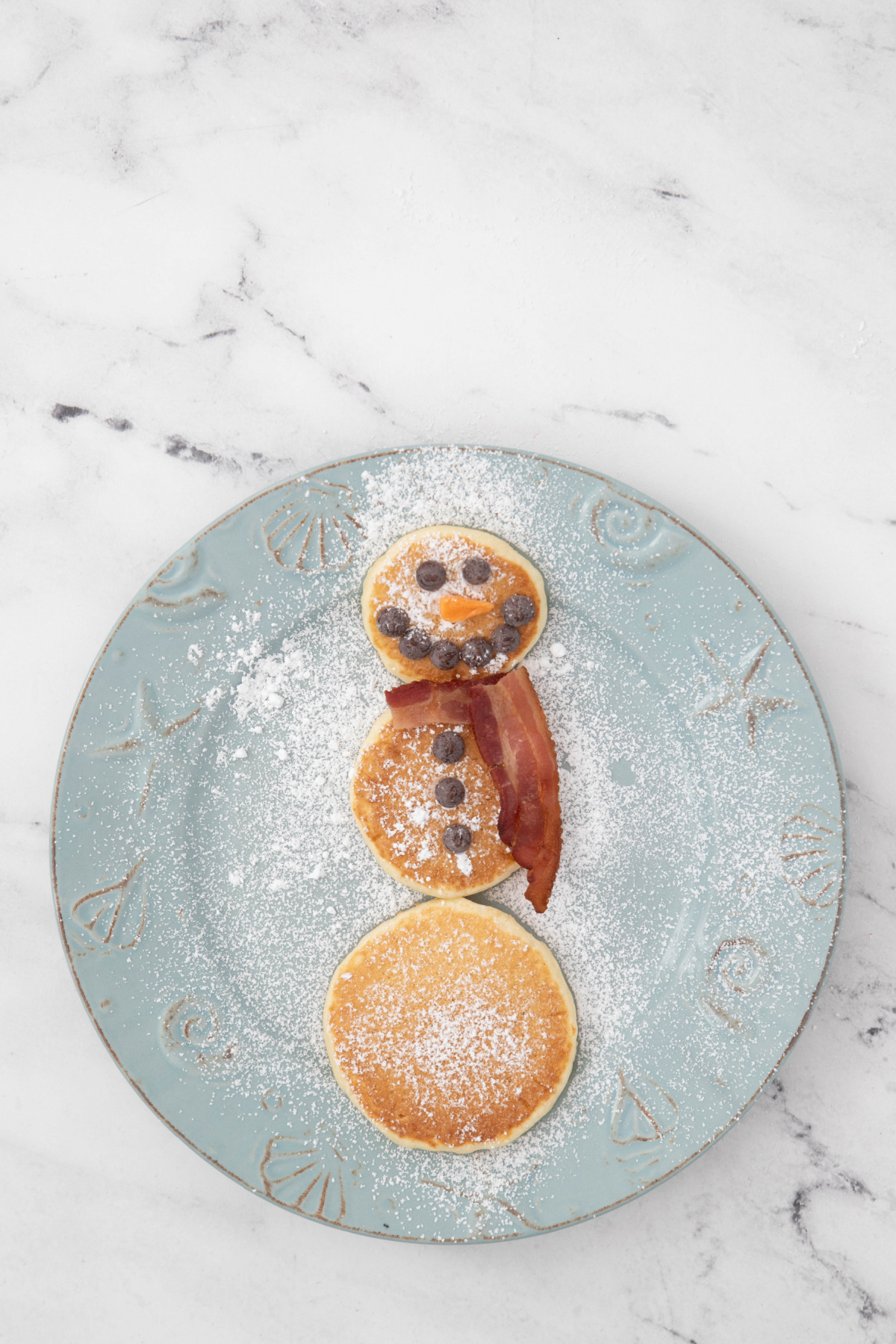 Snowman pancakes with bacon scarf added on a plate with powdered sugar.