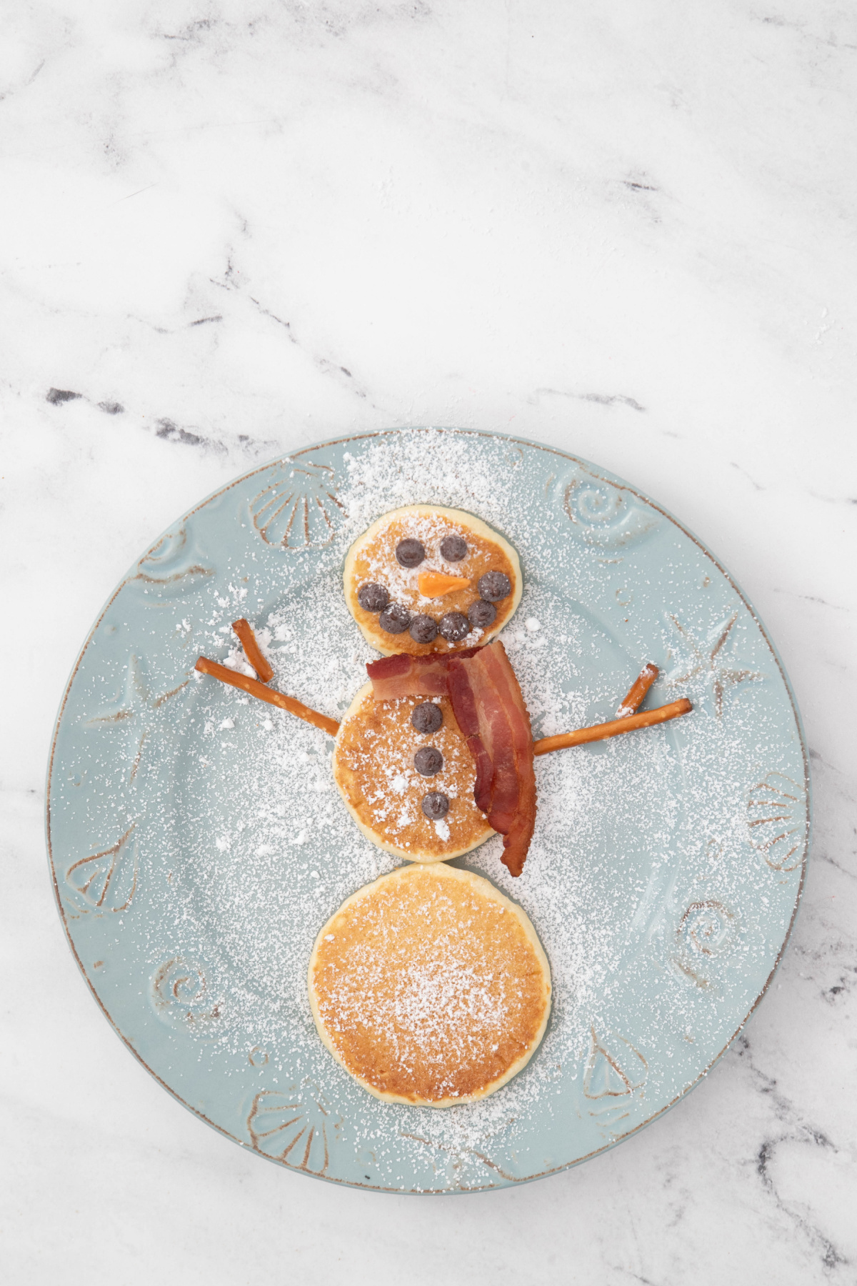 Snowman pancakes with pretzel arms added on a plate with powdered sugar.