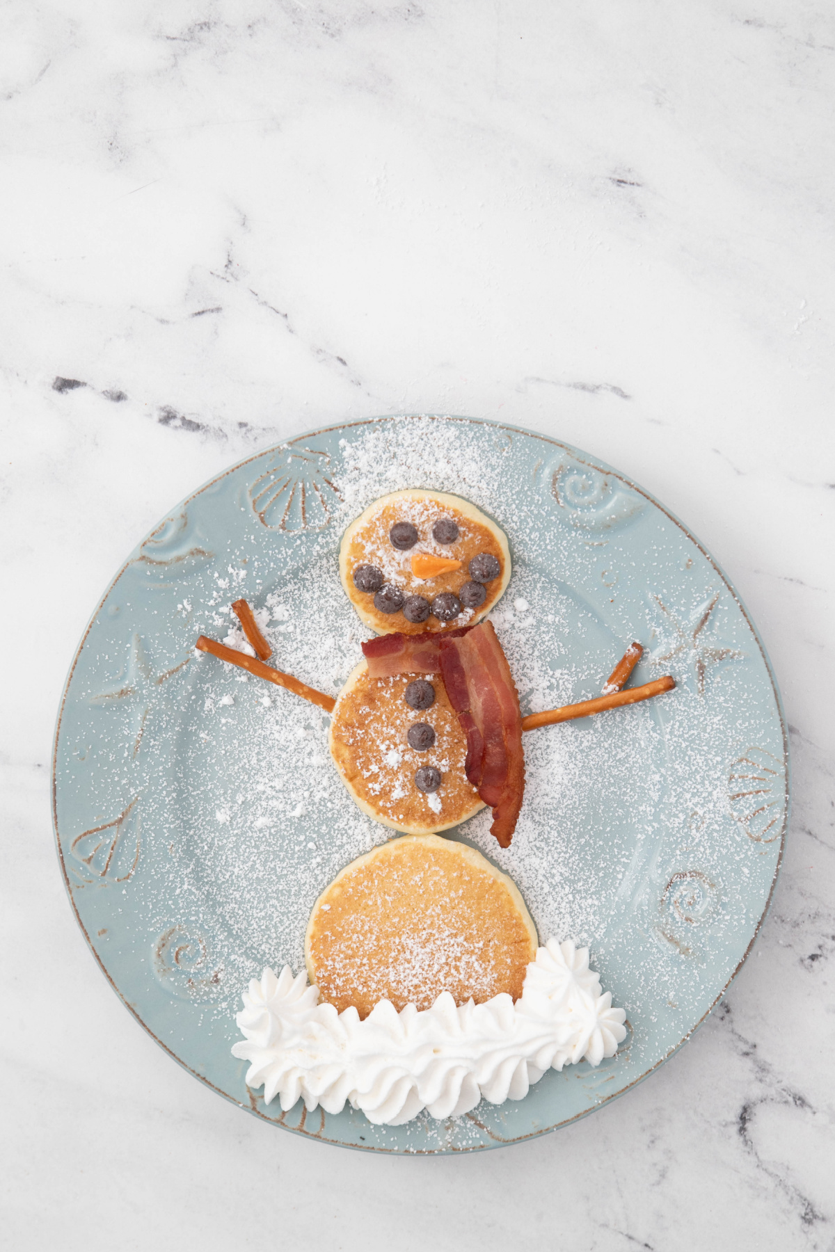 Snowman pancakes with whipped cream snow on a plate with powdered sugar.
