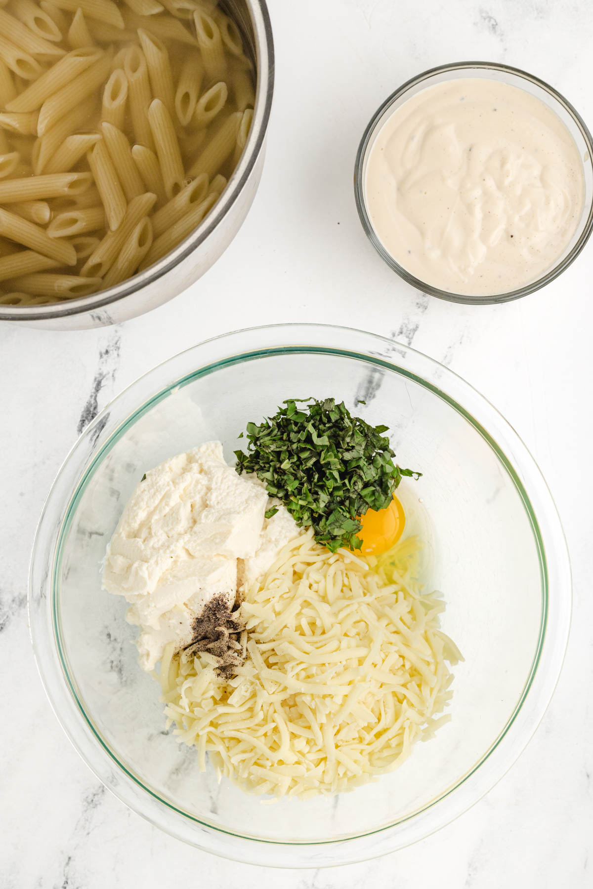 A bowl of cheeses, egg and basil next to a bowl of pasta.