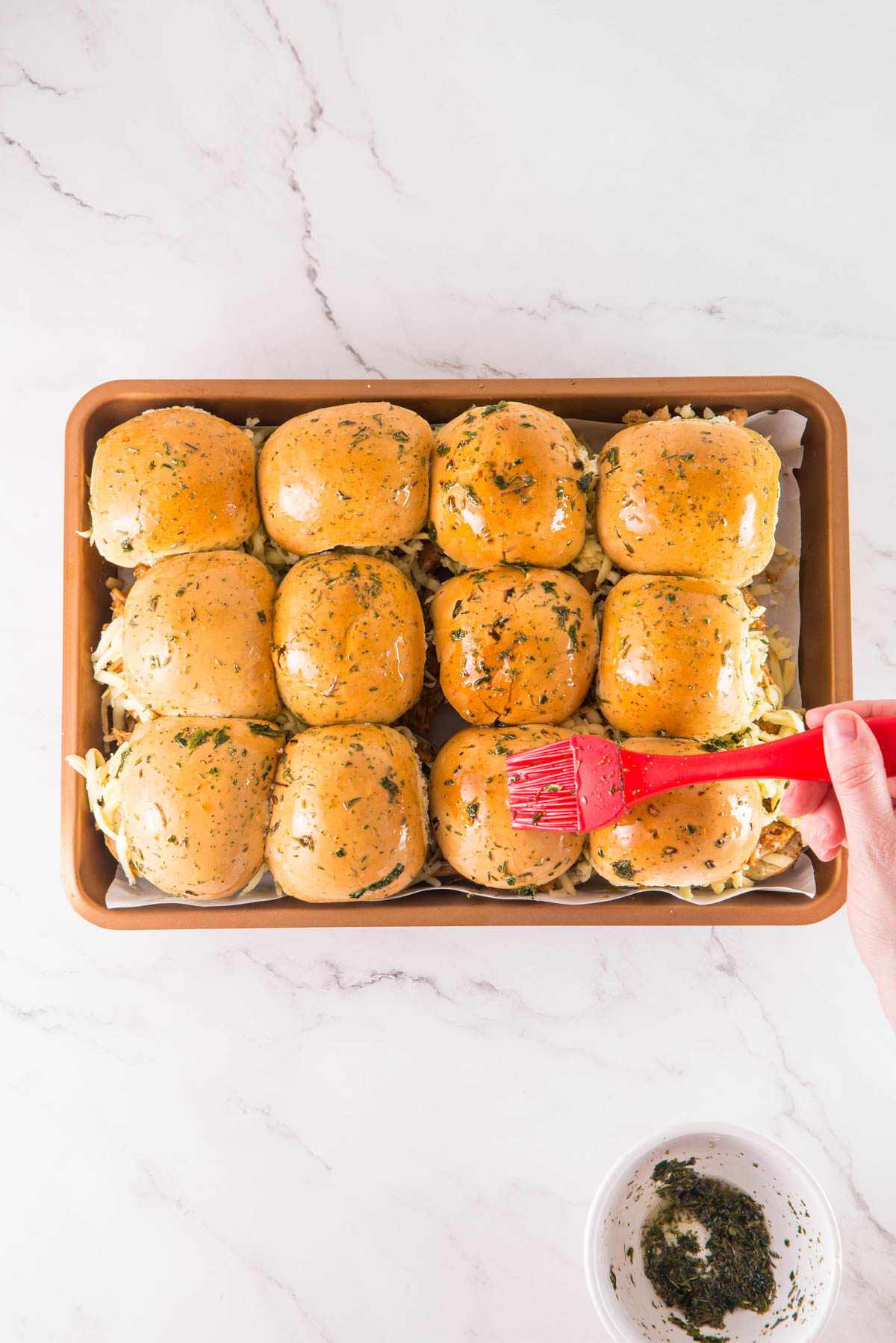 A person is using a red spatula to brush Hawaiian slider rolls with butter and herbs