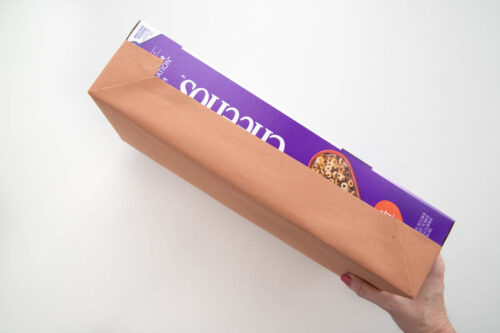 A person holding a cereal box half covered in brown paper in front of a white background.