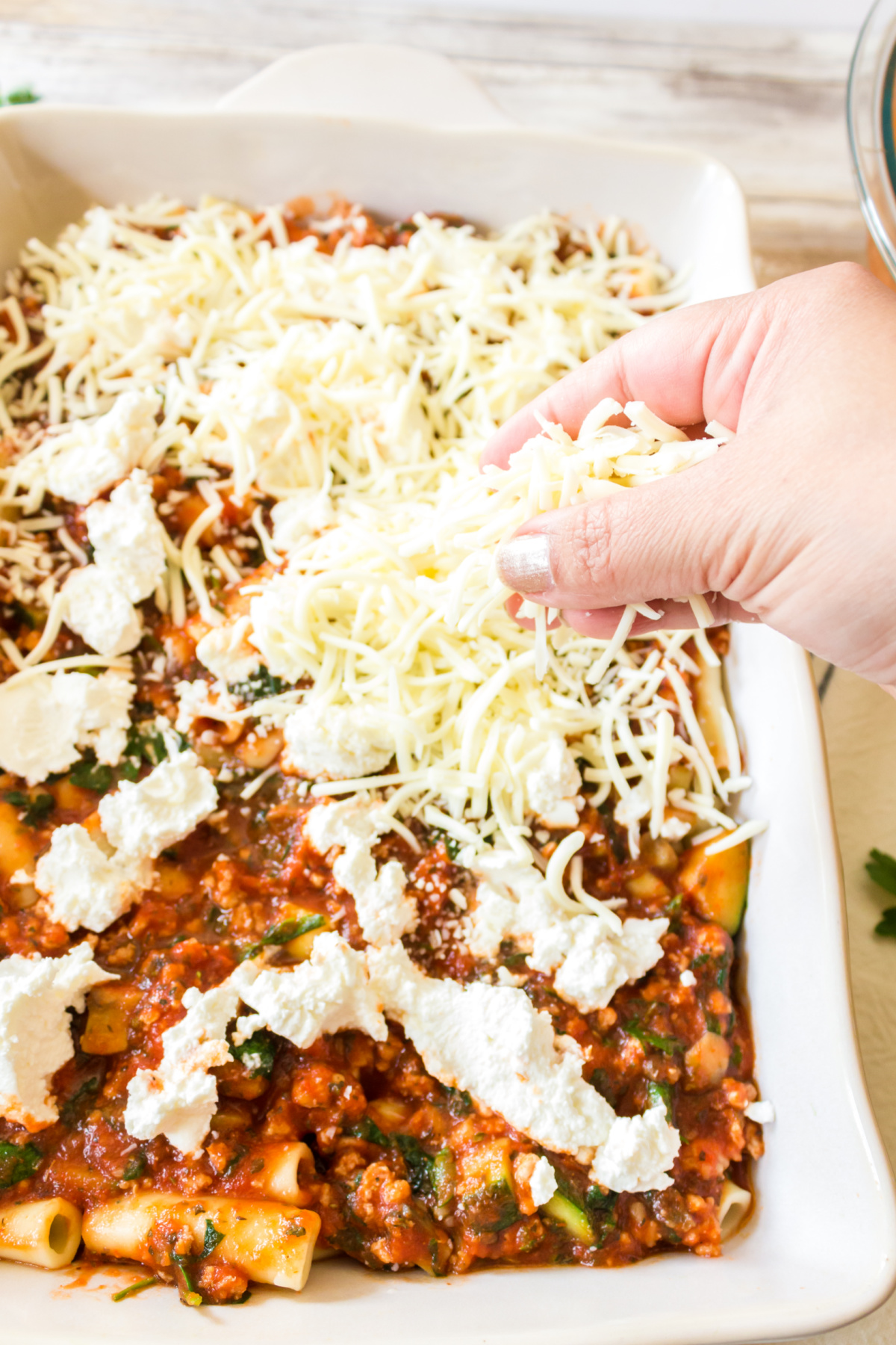 A person's hand sprinkling shredded cheese over a baking dish filled with a pasta dish.
