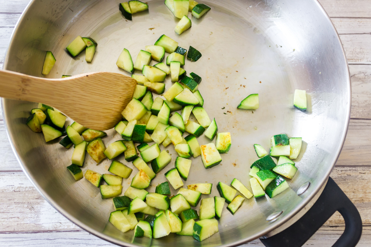 Diced zucchini being sautéed in a stainless steel pan with a wooden spatula.