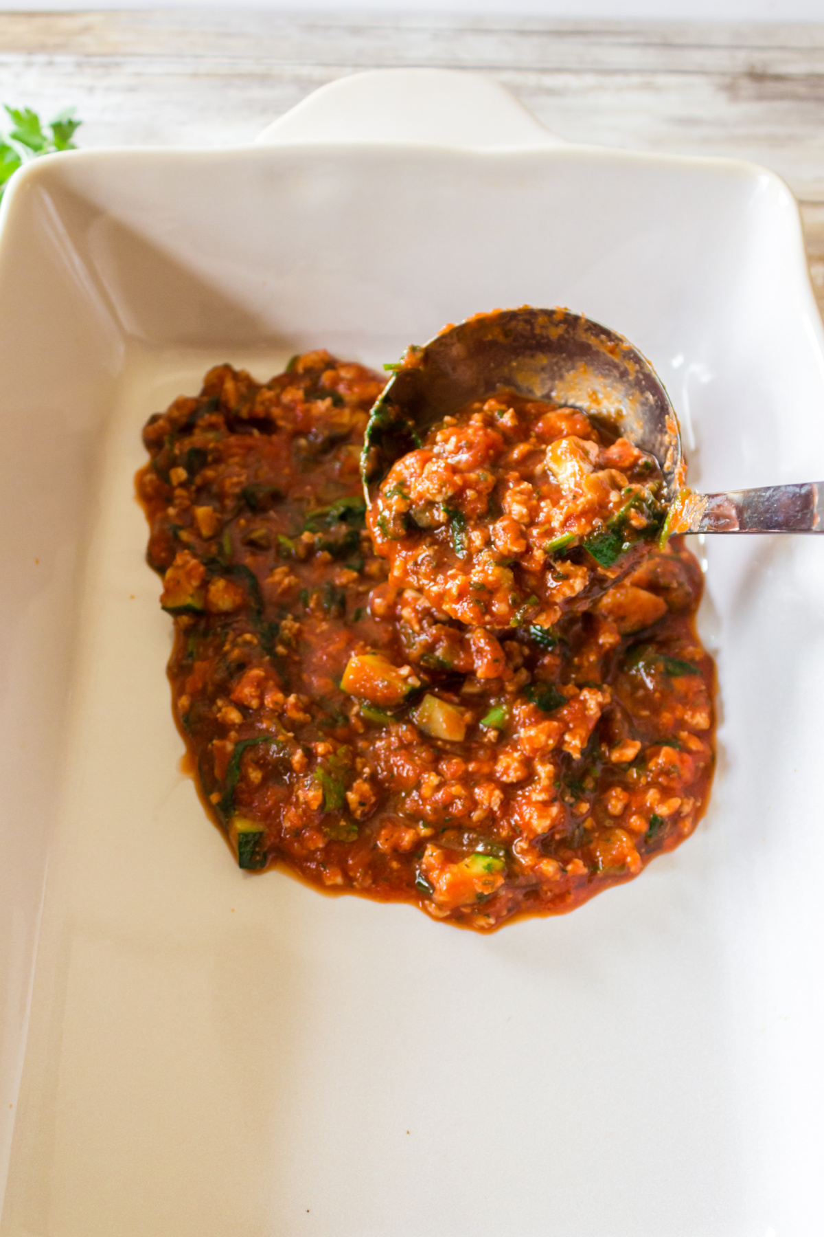 A ladle serving a portion of chunky tomato-based vegetable stew into a white baking dish