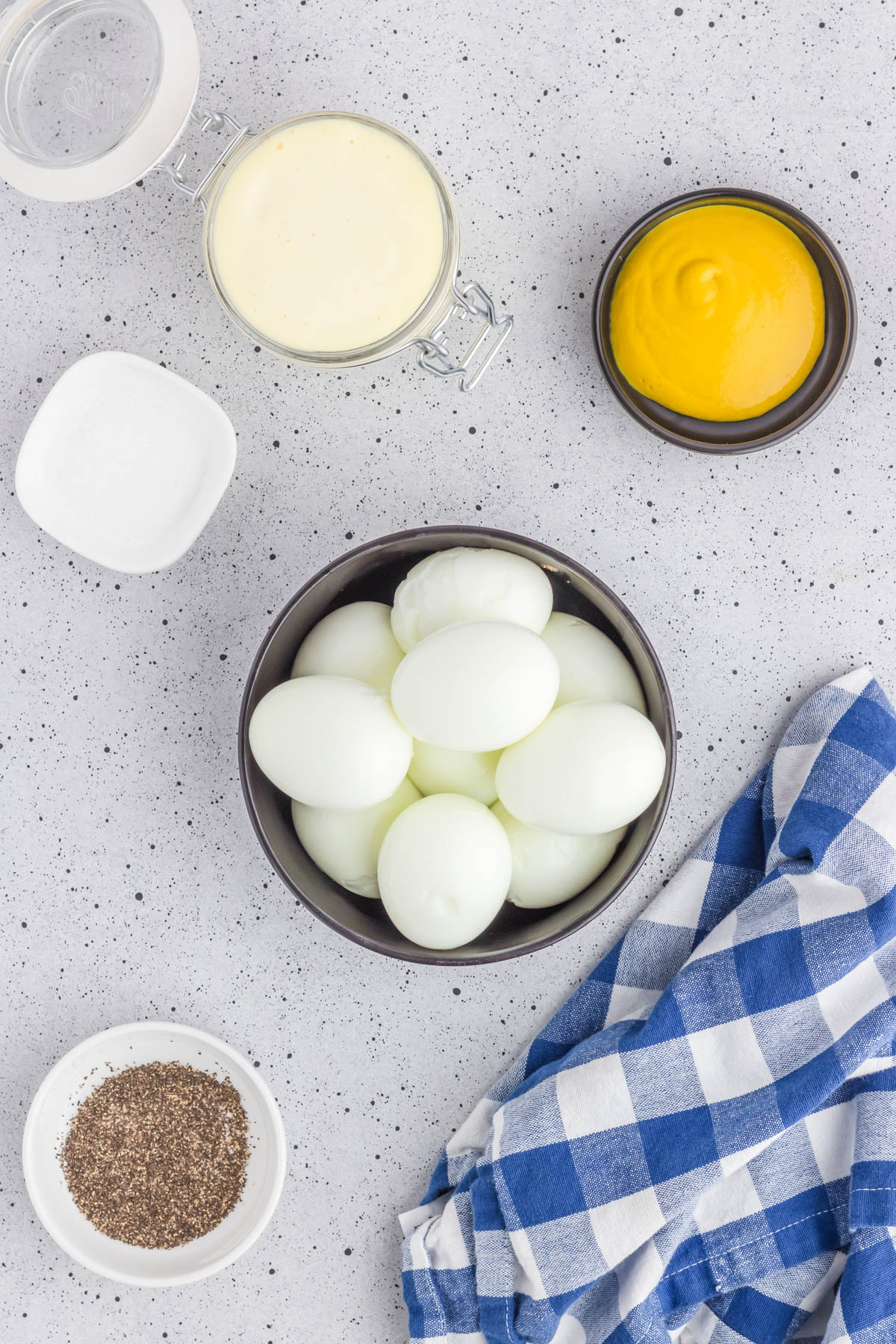 Ingredients for making egg salad including hard-boiled eggs, mayonnaise, mustard, salt, and pepper on a kitchen counter.