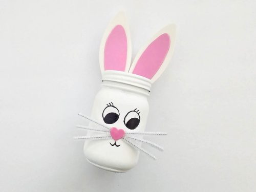 Crafted bunny decoration made from a mason jar with pink interior ears, drawn-on eyes, a pink nose, and string whiskers on a white background.