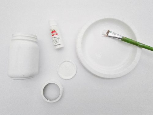 An assortment of art supplies including a jar of white paint, a bottle of glue, a plastic plate, and a green paintbrush, arranged on a white surface.