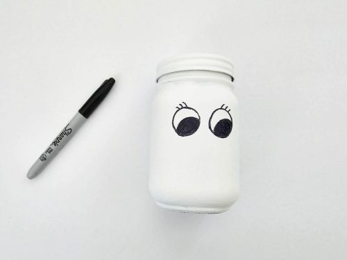 A white jar with cartoon eyes drawn on it, placed next to a black marker on a white background.