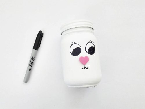 A white jar with a cute face drawn on it next to a black marker on a plain background.