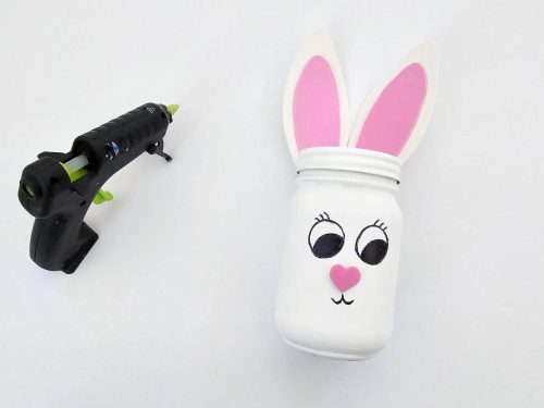 A mason jar with bunny ears and a face next to a glue gun on a white background.
