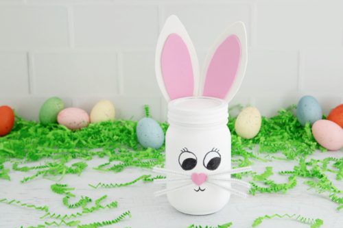 A jar painted white with pink bunny ears, a drawn face, and a whisker decoration, surrounded by colorful easter eggs and green shredded paper.