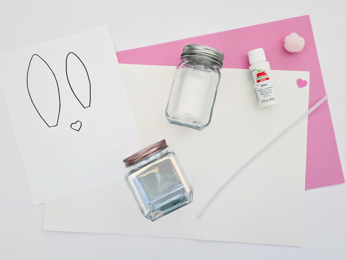 Diy craft supplies including paper with bunny ears drawing, a glass jar, glue, and paintbrush on white and pink craft foam