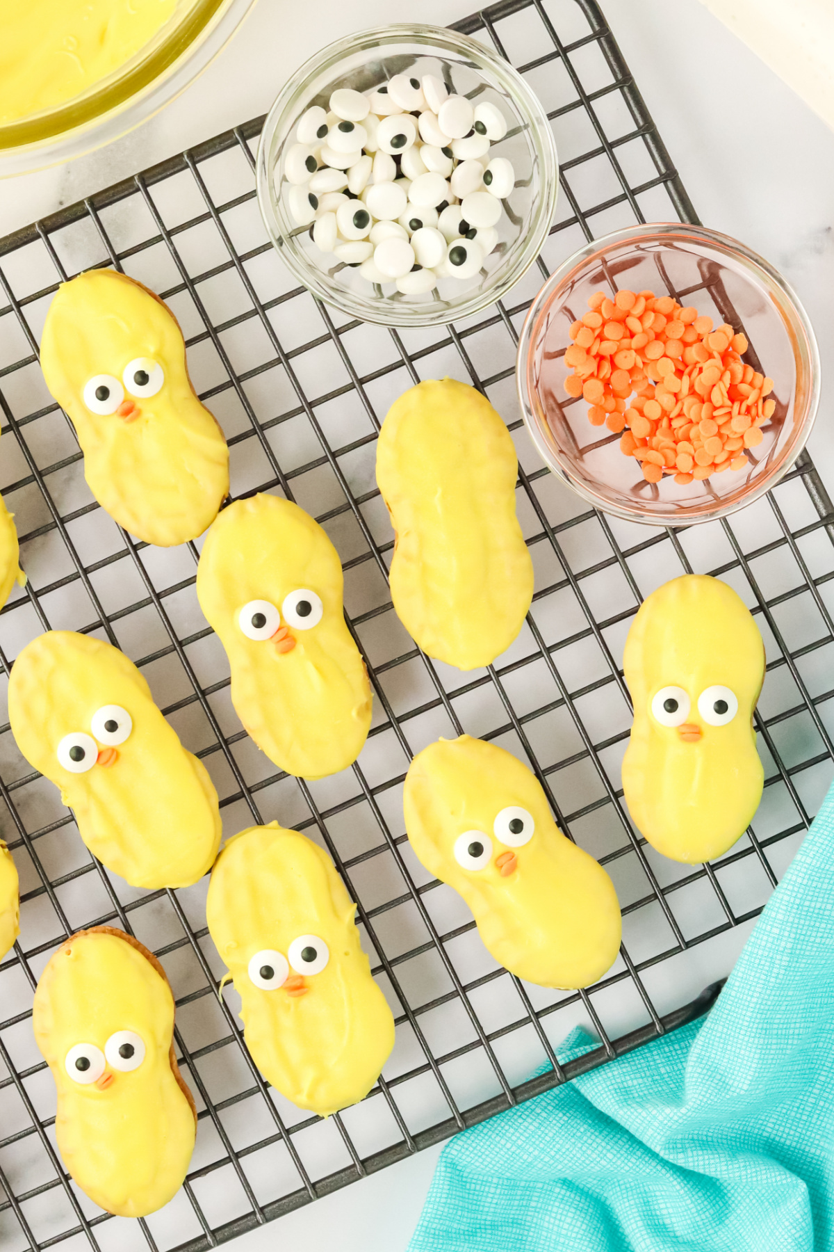 Decorated chick-shaped cookies with candy eyes on a cooling rack, with ingredients nearby.