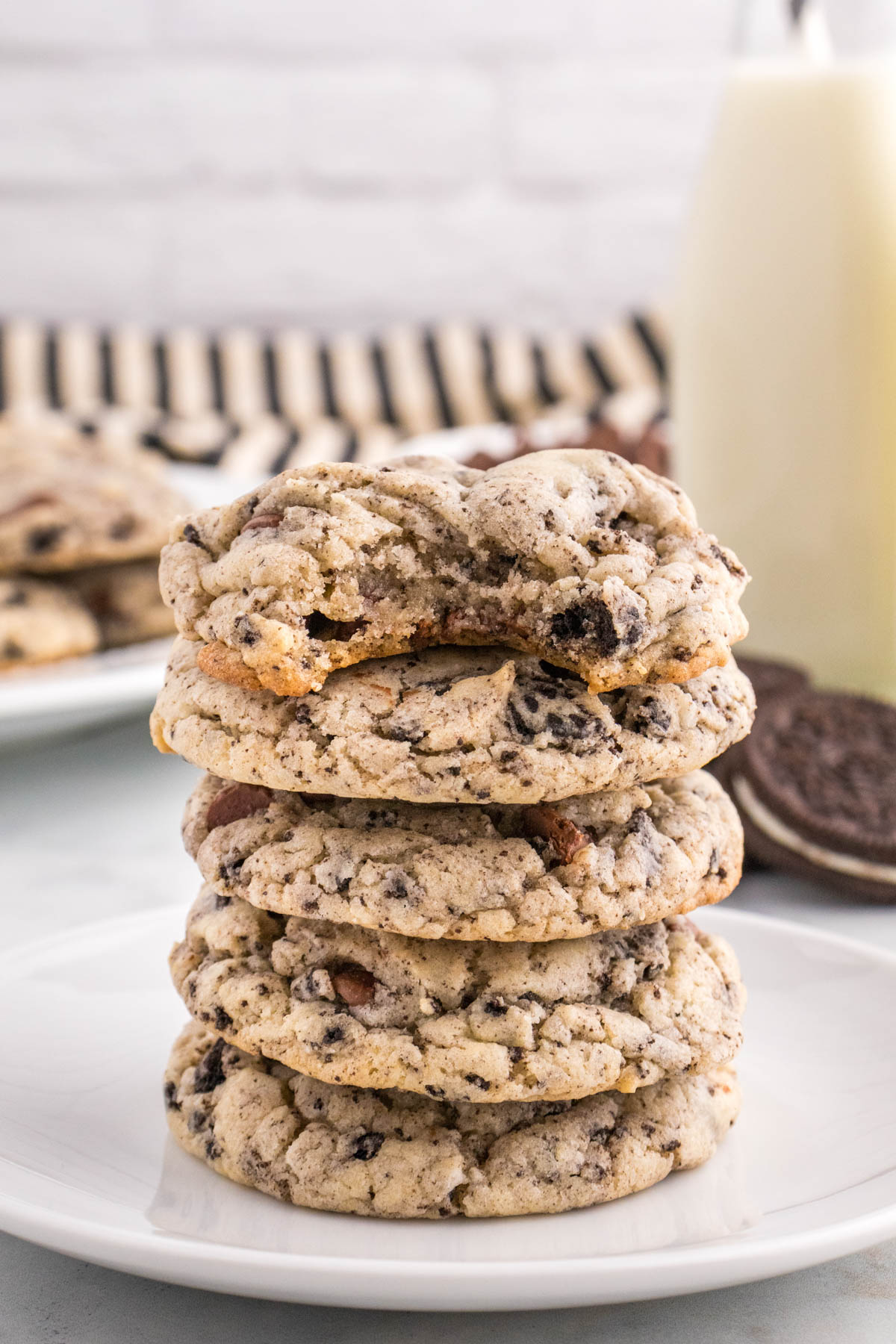 A stack of chocolate chip cookies on a white plate with a glass of milk in the background.