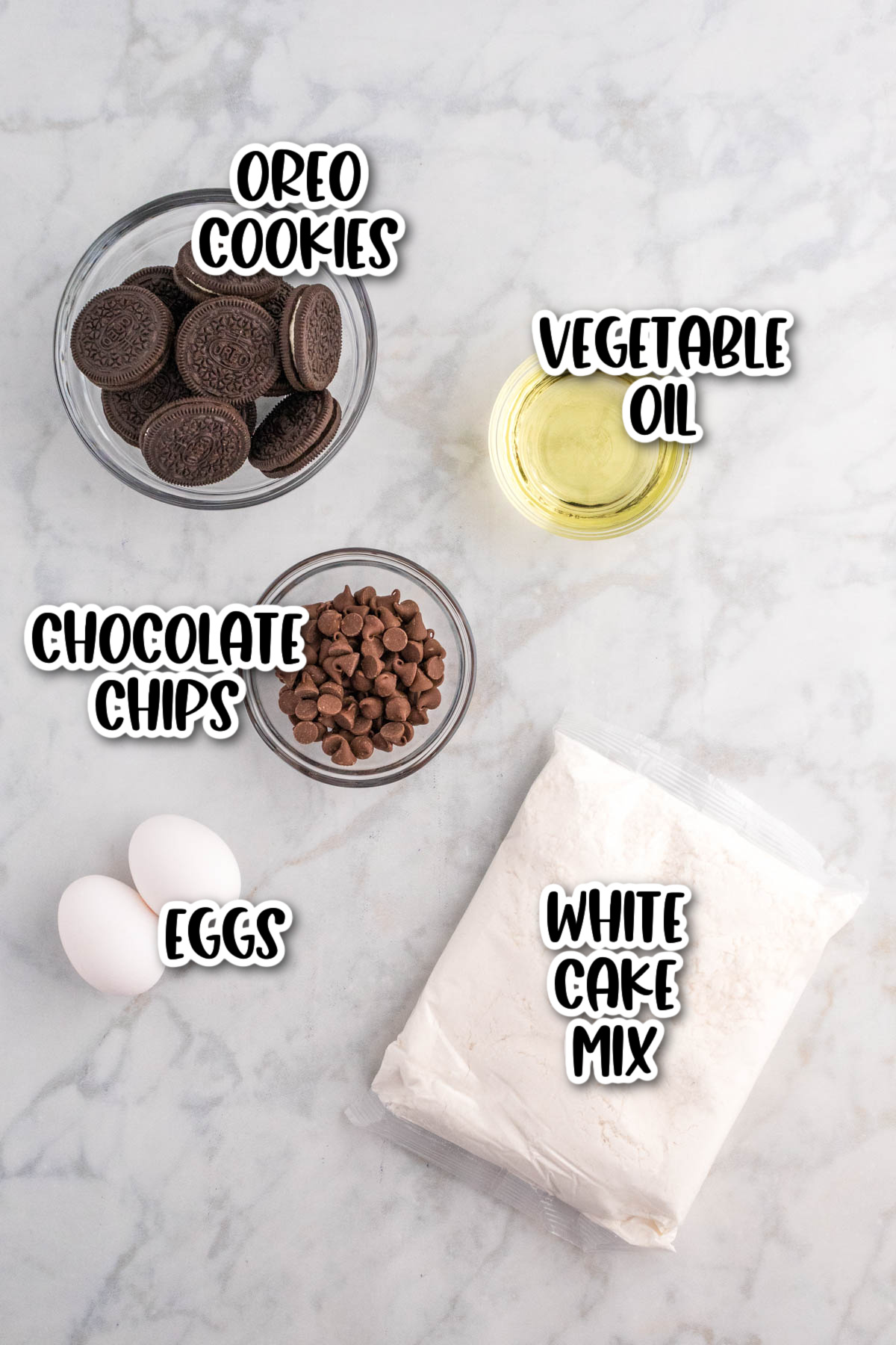 Ingredients for baking laid out on a marble surface, labeled: oreo cookies, vegetable oil, chocolate chips, eggs, and white cake mix.