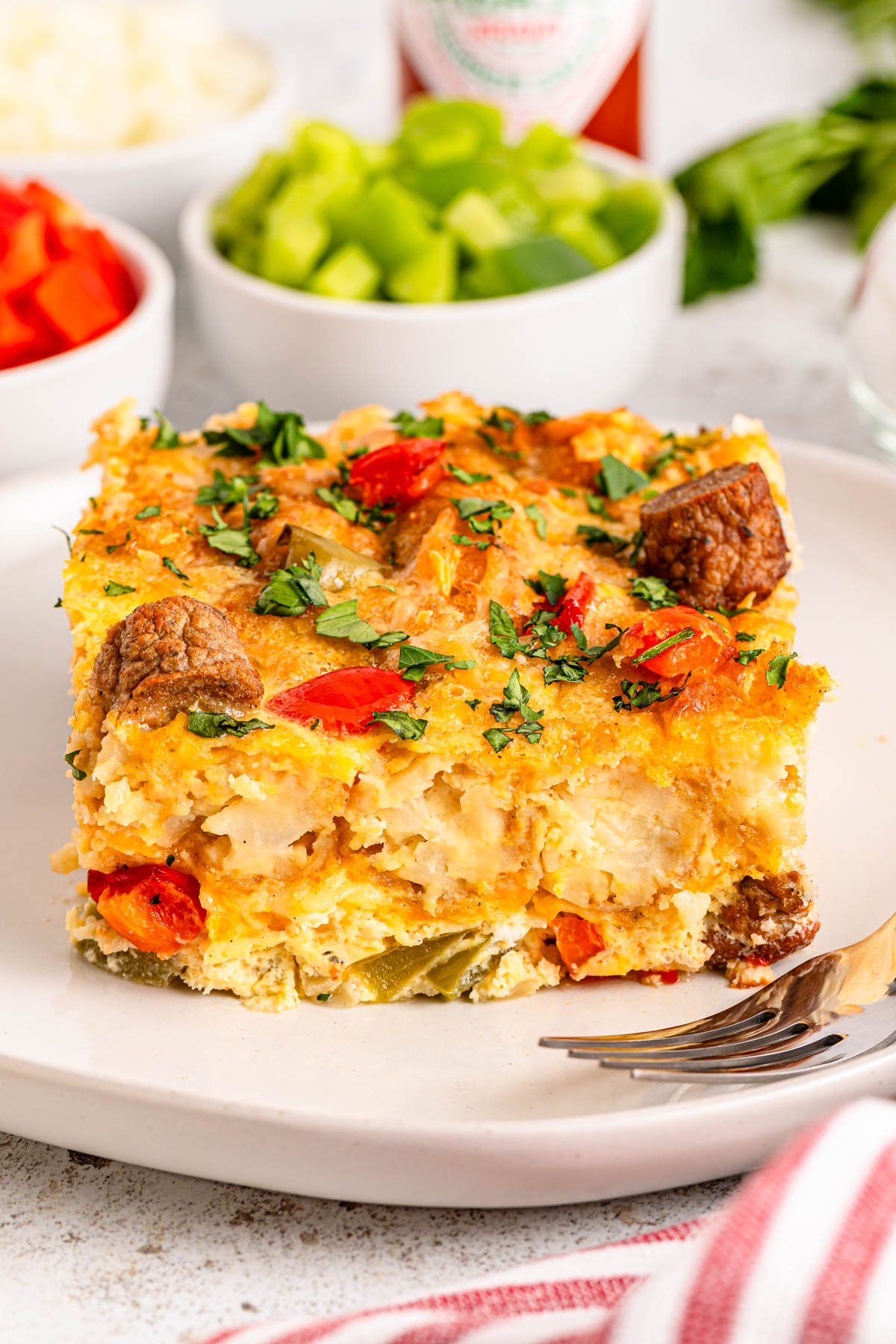 A slice of a breakfast casserole with vegetables and sausage on a plate, garnished with fresh herbs.