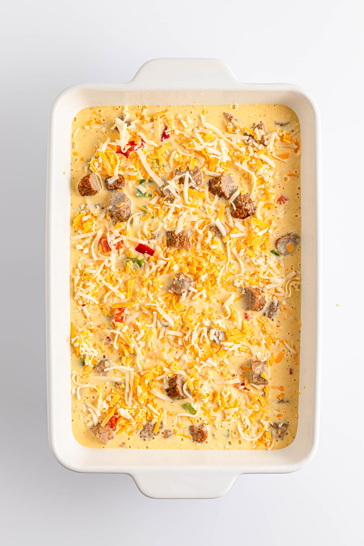 Uncooked casserole dish with shredded cheese and mixed ingredients on a white background.