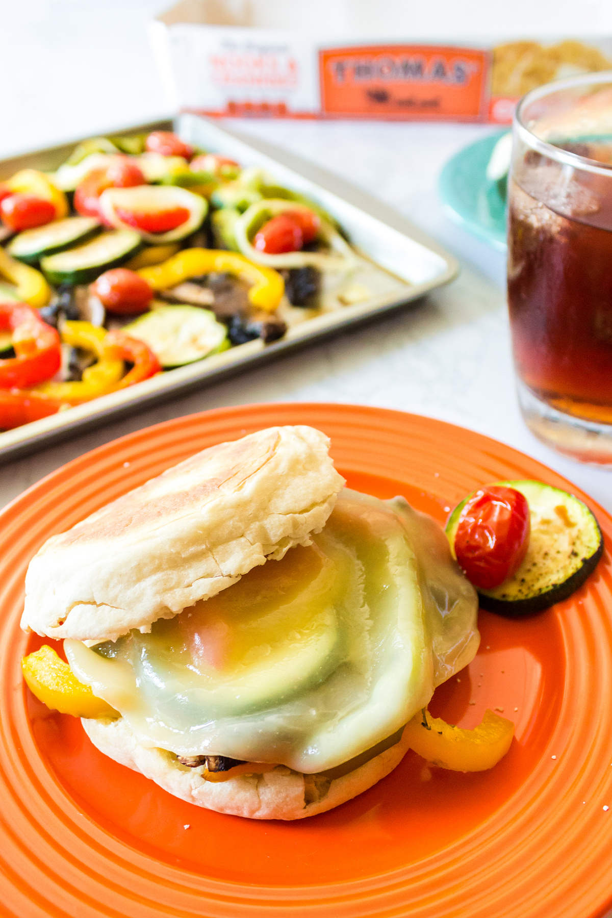 A sandwich with veggies and cheese on an English muffin, served on an orange plate with a side of roasted vegetables and a cold beverage.