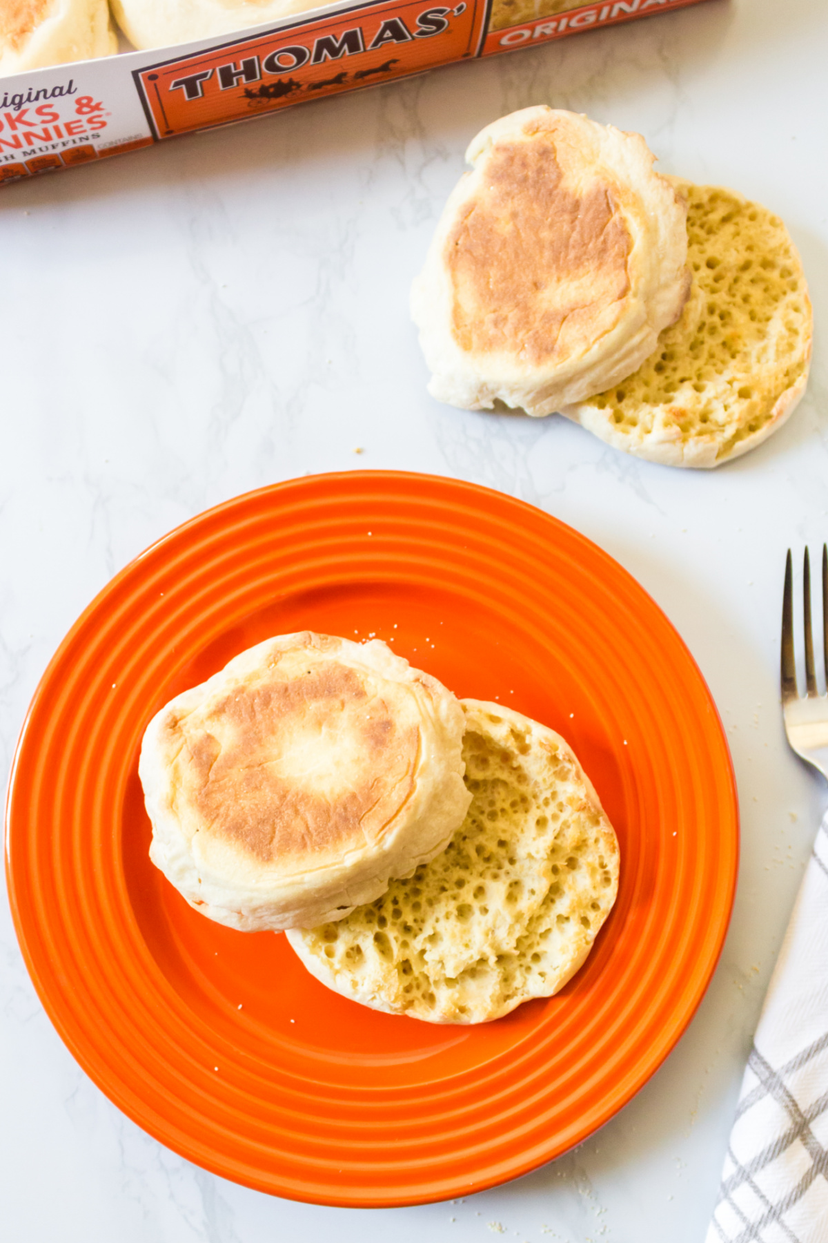 Toasted english muffins on an orange plate with a fork and a pack of thomas' english muffins in the background.