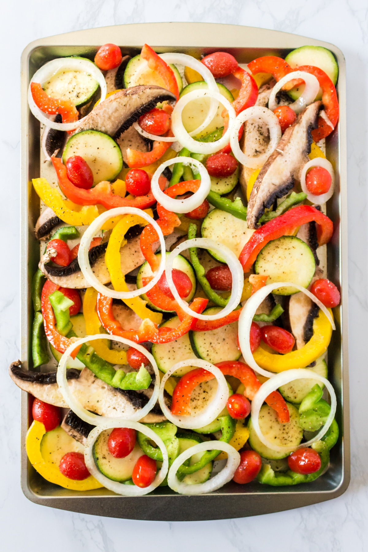An assortment of raw, sliced vegetables seasoned with pepper on a baking sheet, ready to be roasted.