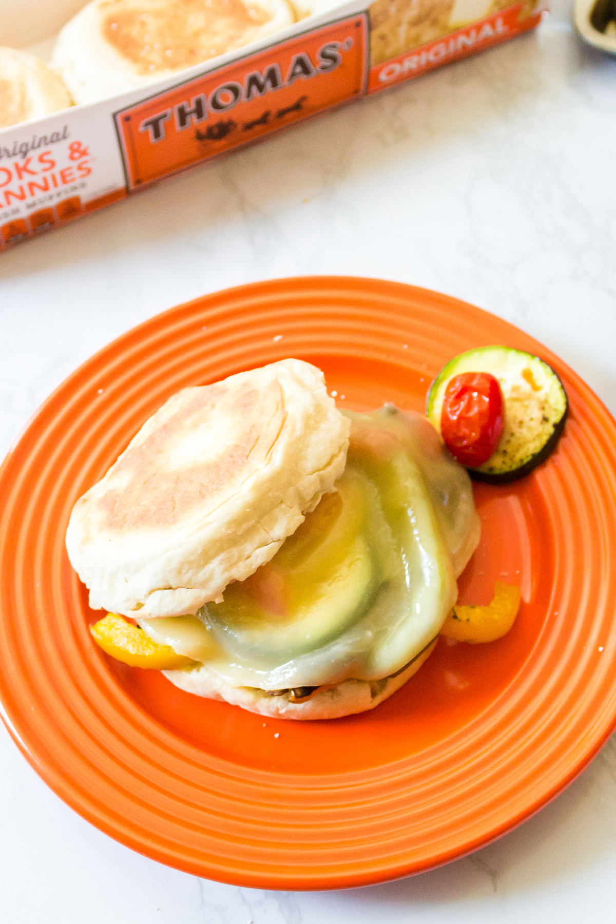 A veggie sandwich with melted cheese on a English muffin, served on an orange plate with a side of cherry tomatoes and zucchini slices.