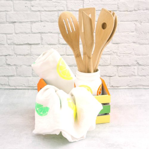 Wooden kitchen utensils in a colorful holder with white towels printed with green lemons, set against a white brick background, perfect for Mother's Day crafts.