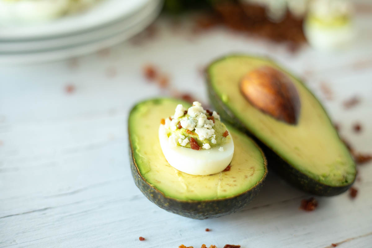 A halved avocado with one half filled with a boiled egg topped with crumbled cheese, on a white surface with scattered bacon