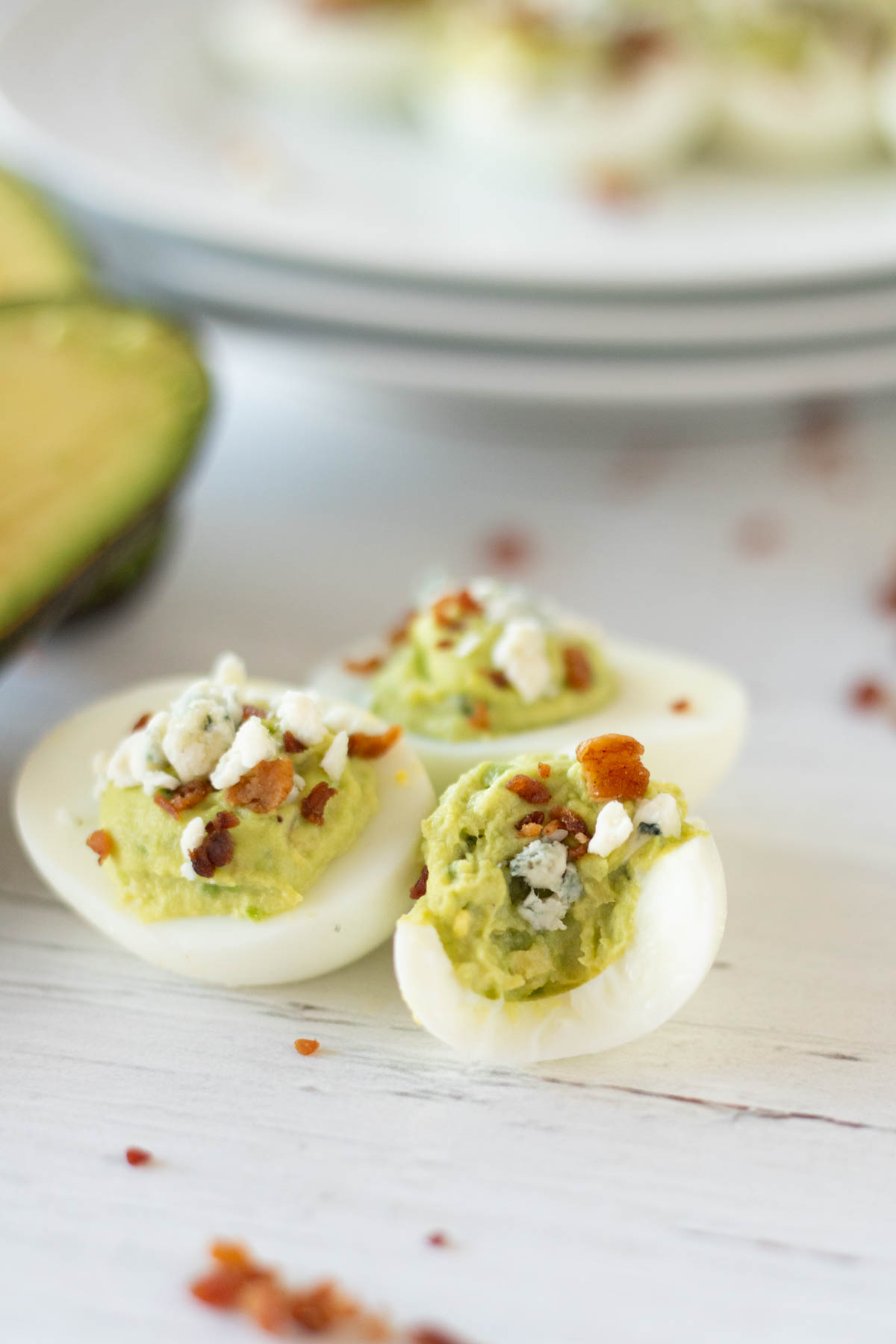 Avocado deviled eggs garnished with crumbled cheese and bacon on a white surface.