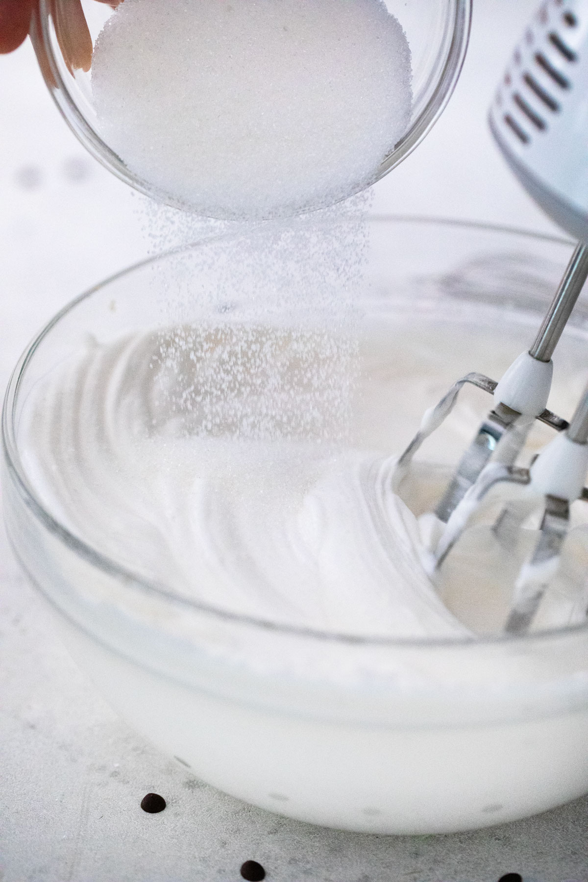 Sugar being poured into egg white mixture in a glass bowl with an electric mixer in action.