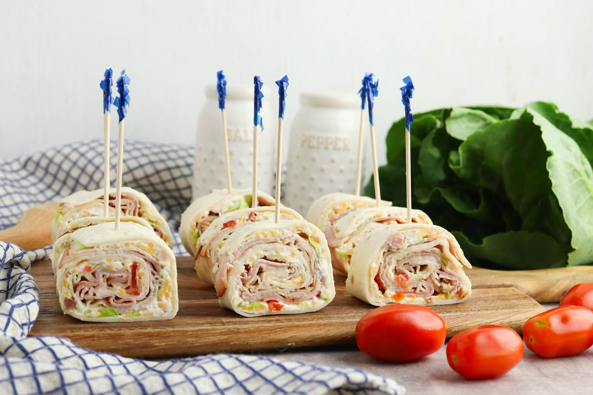 A wooden board with turkey and cheese roll-ups, garnished with toothpicks, accompanied by tomatoes and lettuce, against a kitchen backdrop.