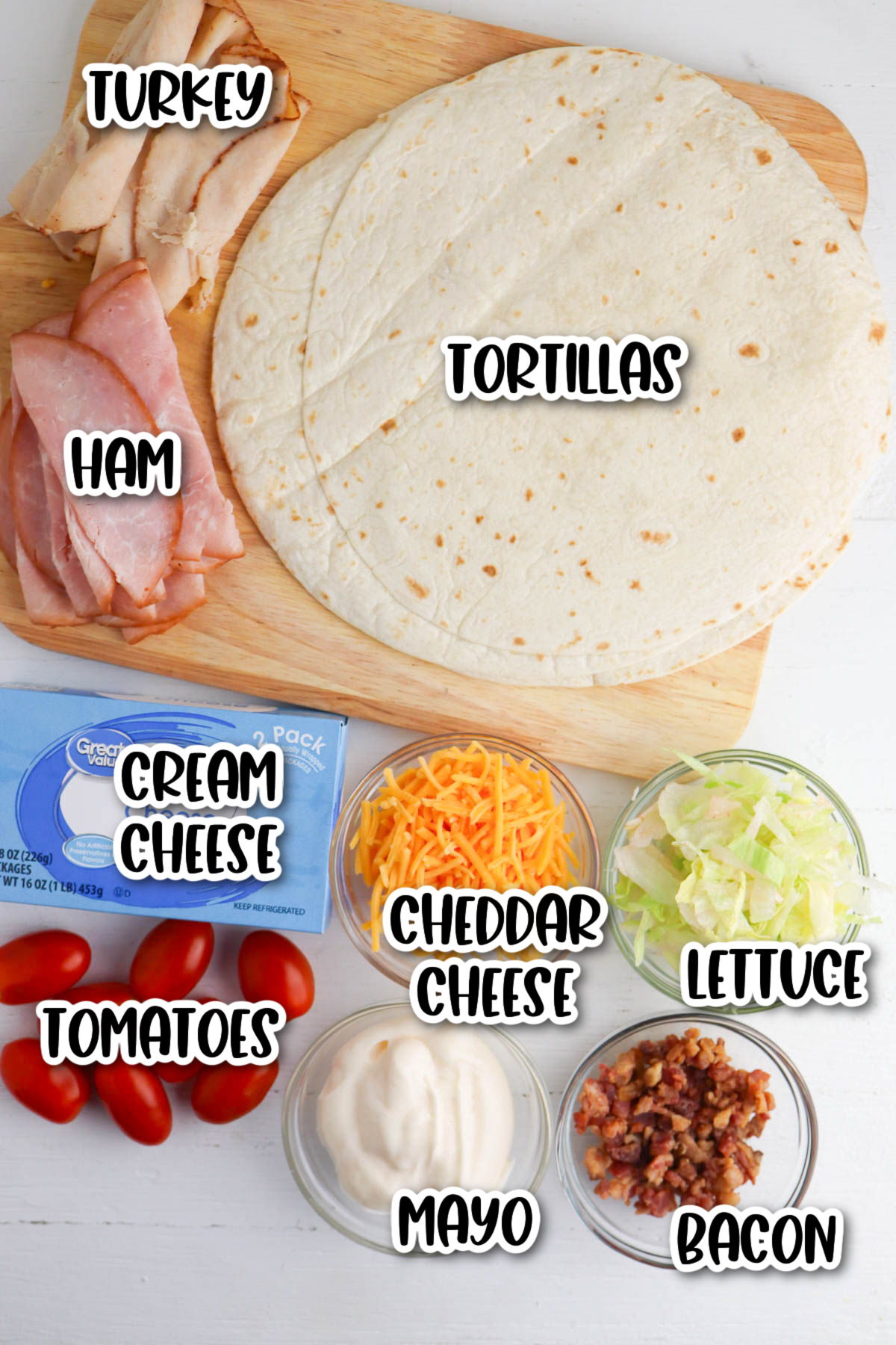 Ingredients laid out for making wraps, including tortillas, turkey, ham, cream cheese, tomatoes, cheddar cheese, lettuce, mayo, and bacon.