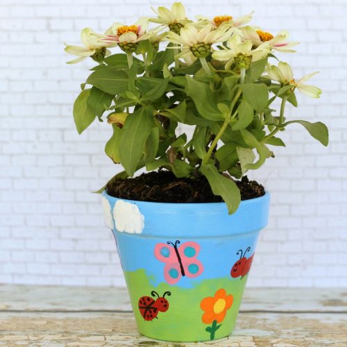 A potted plant with white flowers in a blue pot decorated with colorful butterfly and ladybug designs, ideal for Mother's Day crafts, set against a white brick background.