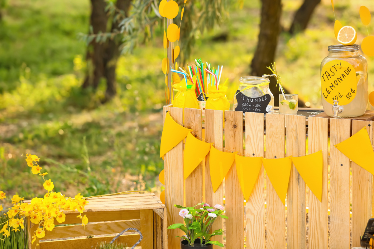 A lemonade stand decorated with yellow bunting in a sunny garden, featuring jars of lemonade, colorful straws, and fresh flowers.