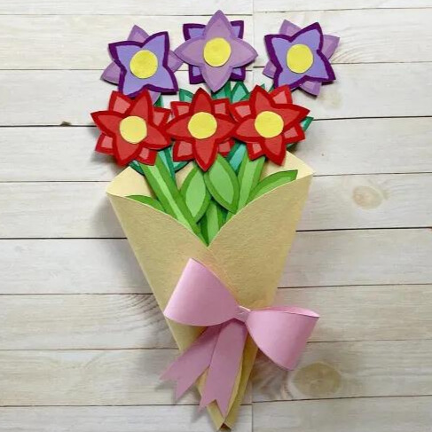 A handmade bouquet of colorful paper flowers, featuring red and purple blooms with green stems, wrapped in a yellow paper cone tied with a pink ribbon.