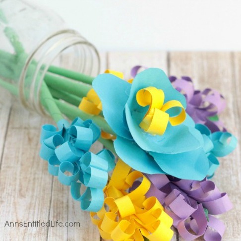 A glass jar tipped over with a bouquet of colorful paper flowers spilling out, featuring blue, yellow, and purple flowers.